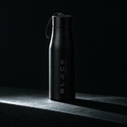 The Black Edition stainless bottle in matte black, standing on a concrete ledge in hard side light, engraved wordmark running vertically up the body.