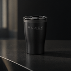The Black Edition travel cup in matte black with the slider lid fitted, on a dark stone benchtop in morning light, engraved wordmark facing the camera.